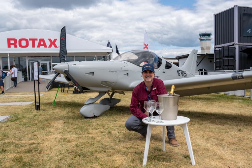 Bernardo Saviñon crouching beside his military-gray Bristell Classic N278BS with a champagne bucket at the Rotax exhibit, EAA AirVenture Oshkosh 2026