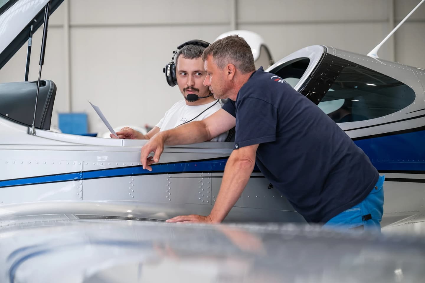 Zwei Männer gehen an einem Bristell-Flugzeug in der Halle ein gedrucktes Dokument durch, einer sitzt mit Flugheadset im offenen Cockpit