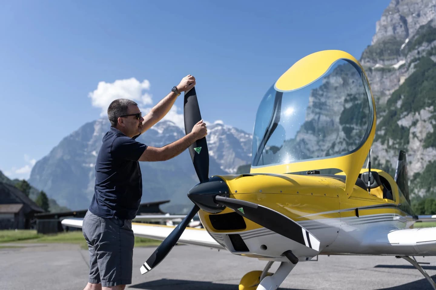 A three-blade MT-Propeller constant-speed propeller on the yellow nose of a Bristell B23, with a person handling one blade against a backdrop of alpine mountains.