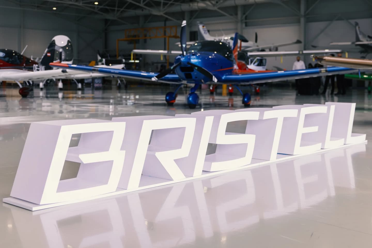 A blue Bristell light aircraft in a bright hangar, behind large free-standing white letters spelling Bristell