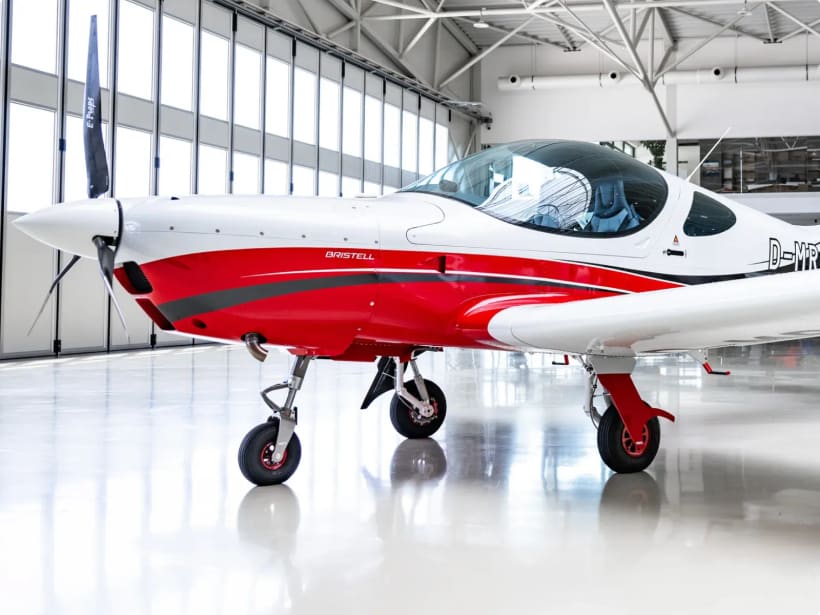 Red-and-white Bristell with retractable landing gear, parked in a hangar.