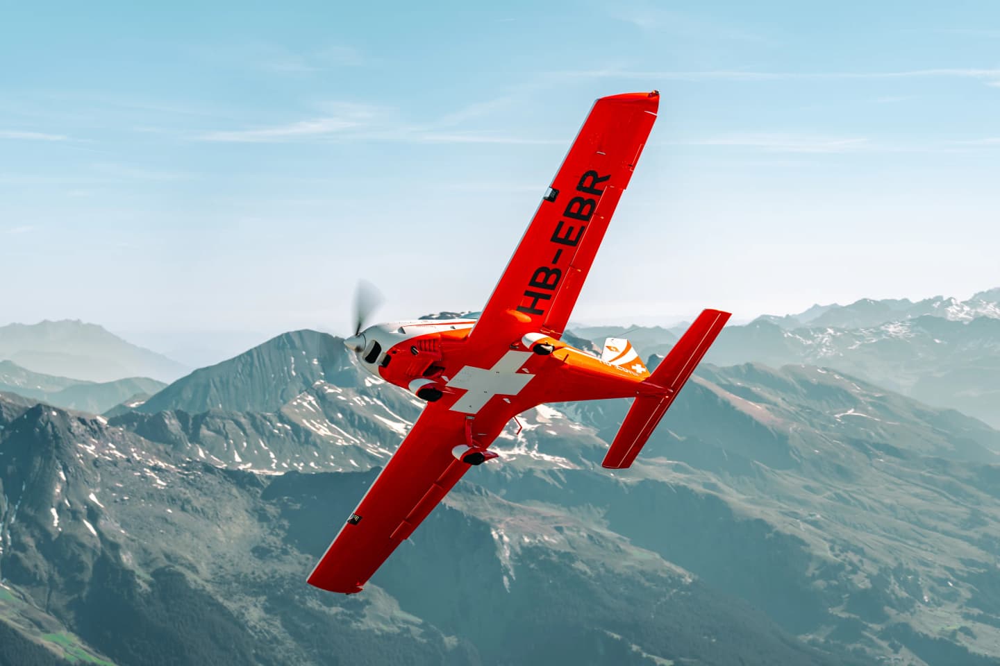 Red Bristell B23 in Swiss livery banking over snow-capped Alpine peaks